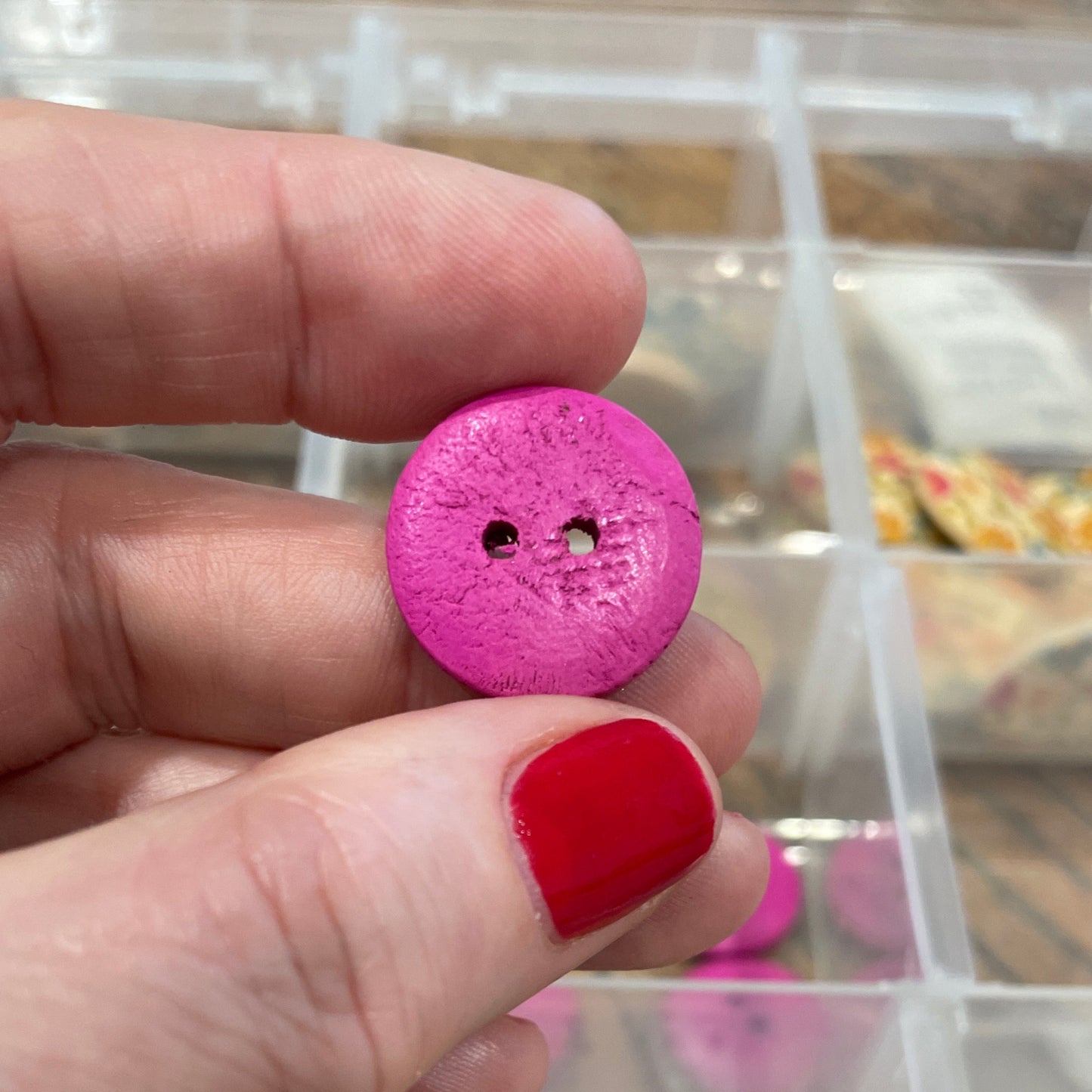 Hand holding a pink button with a container of various buttons in the background