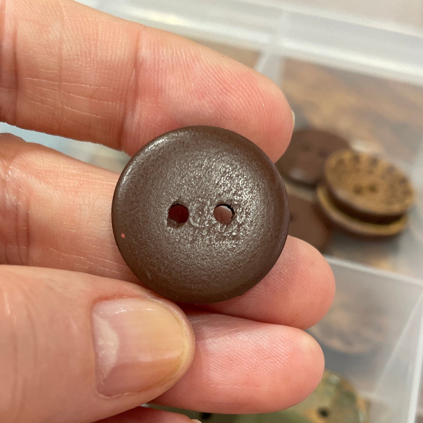 Hand holding a brown button with a container of various buttons in the background