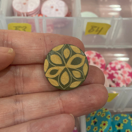 Hand holding a decorative button with a floral design in front of a container of buttons.