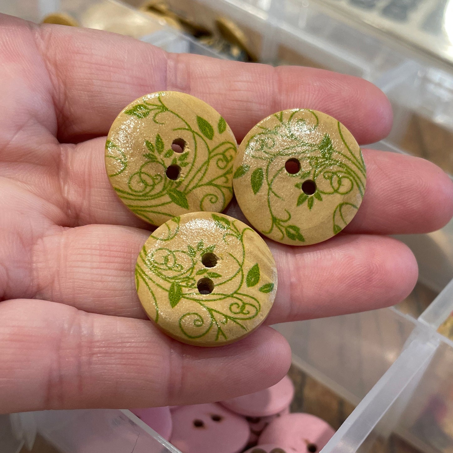Three wooden buttons with green patterns held in a hand, with a container of various buttons in the background.