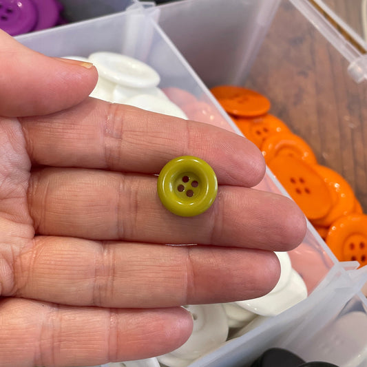 Hand holding a small green button with a container of assorted buttons in the background