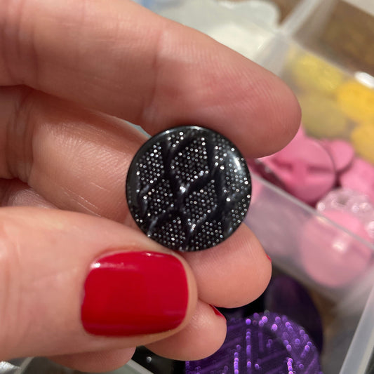 Hand holding a black glittery button with a colorful assortment of buttons in the background.