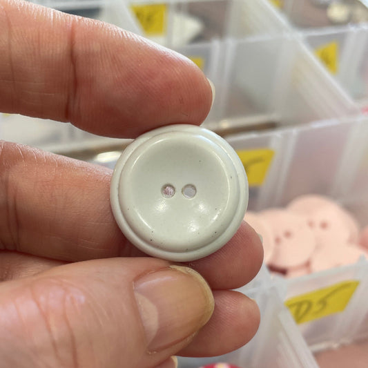 Hand holding a white button with a background of various buttons in a box.