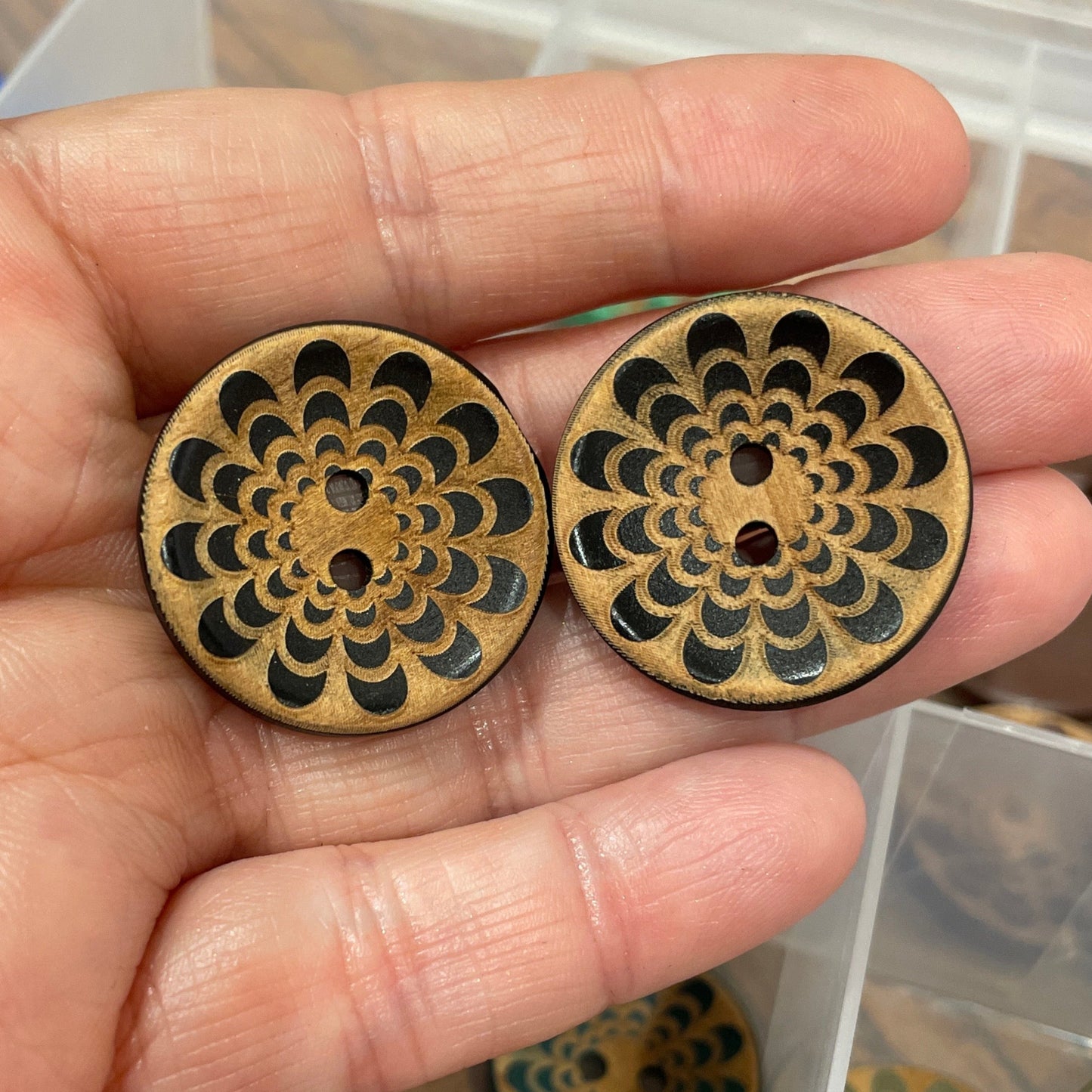 Hand holding two decorative wooden buttons with intricate designs, with a container of similar buttons in the background.