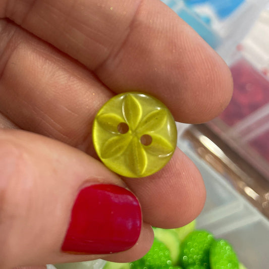 Hand holding a yellow button with a blurred background of green buttons and a clear container.