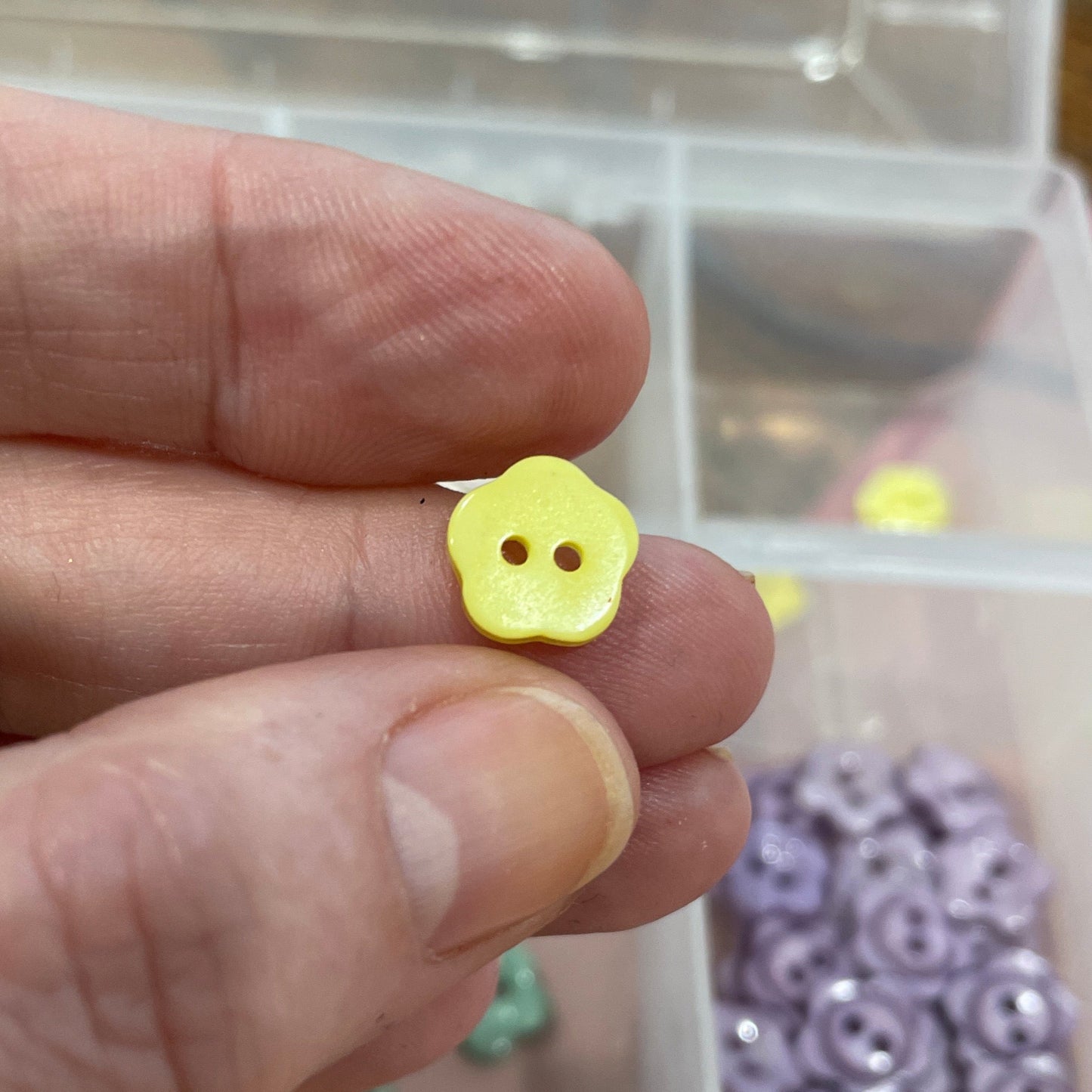 Hand holding a small yellow button with a clear container of buttons in the background