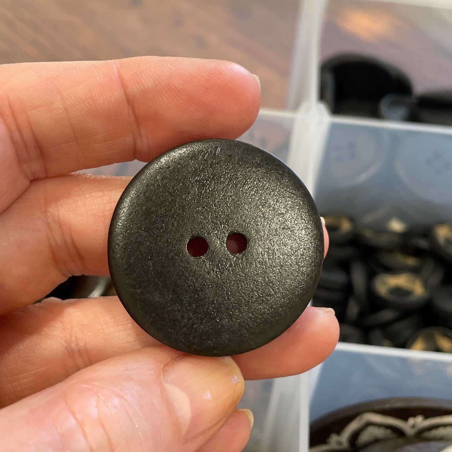 Hand holding a dark brown button with a blurred container of buttons in the background