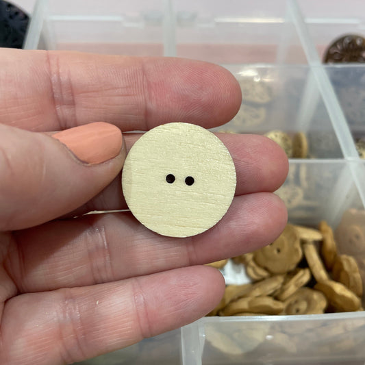 Hand holding a round wooden button with two holes, with a container of buttons in the background.