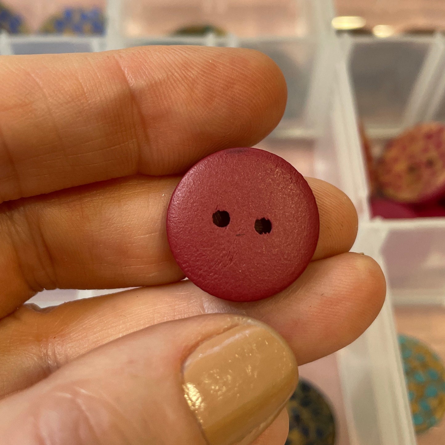 Hand holding a red button with two holes in front of a clear container with colorful buttons.