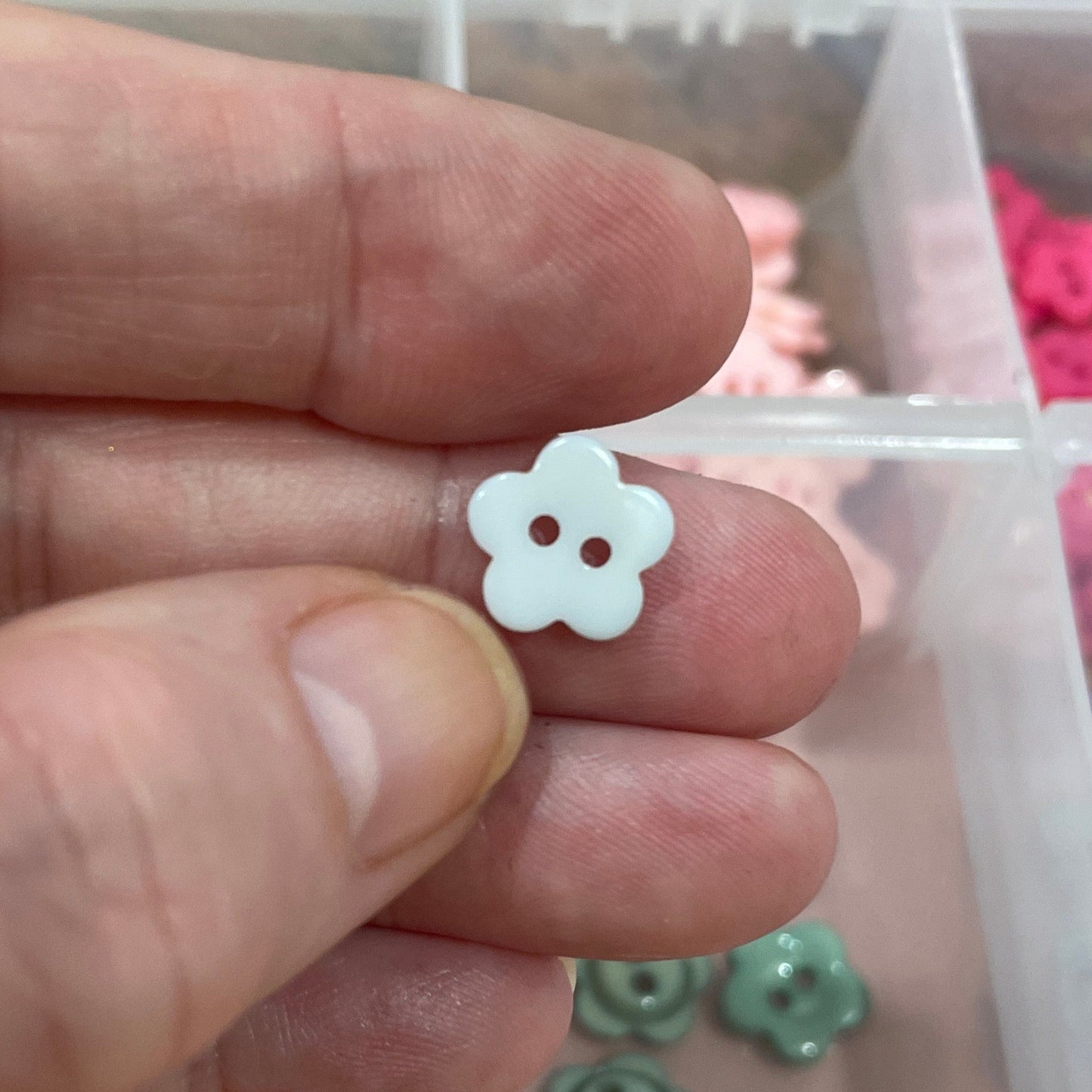 Hand holding a small white flower-shaped object with a container of colorful buttons in the background.