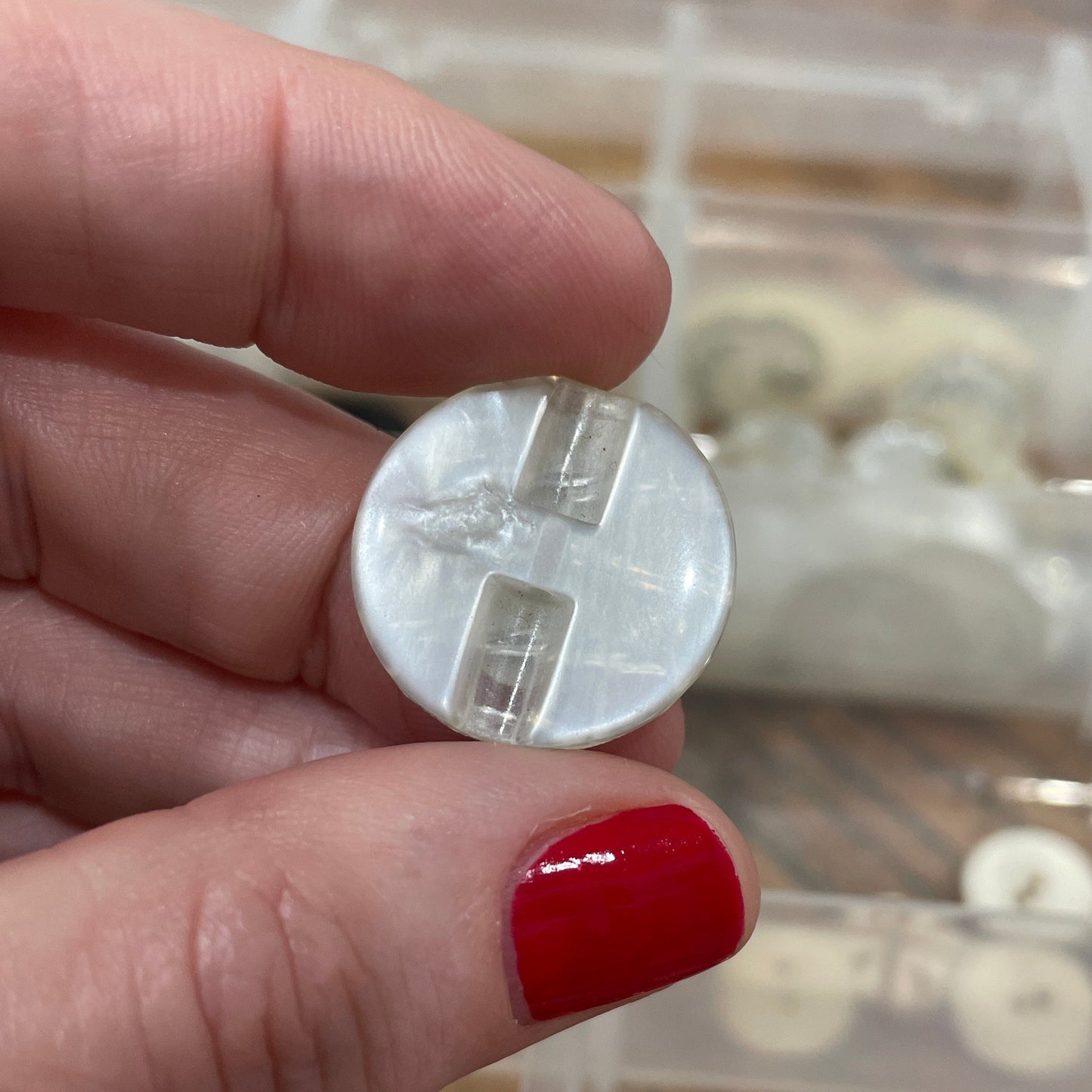 Hand holding a small round object with a clear plastic cover, against a background of buttons in a container.
