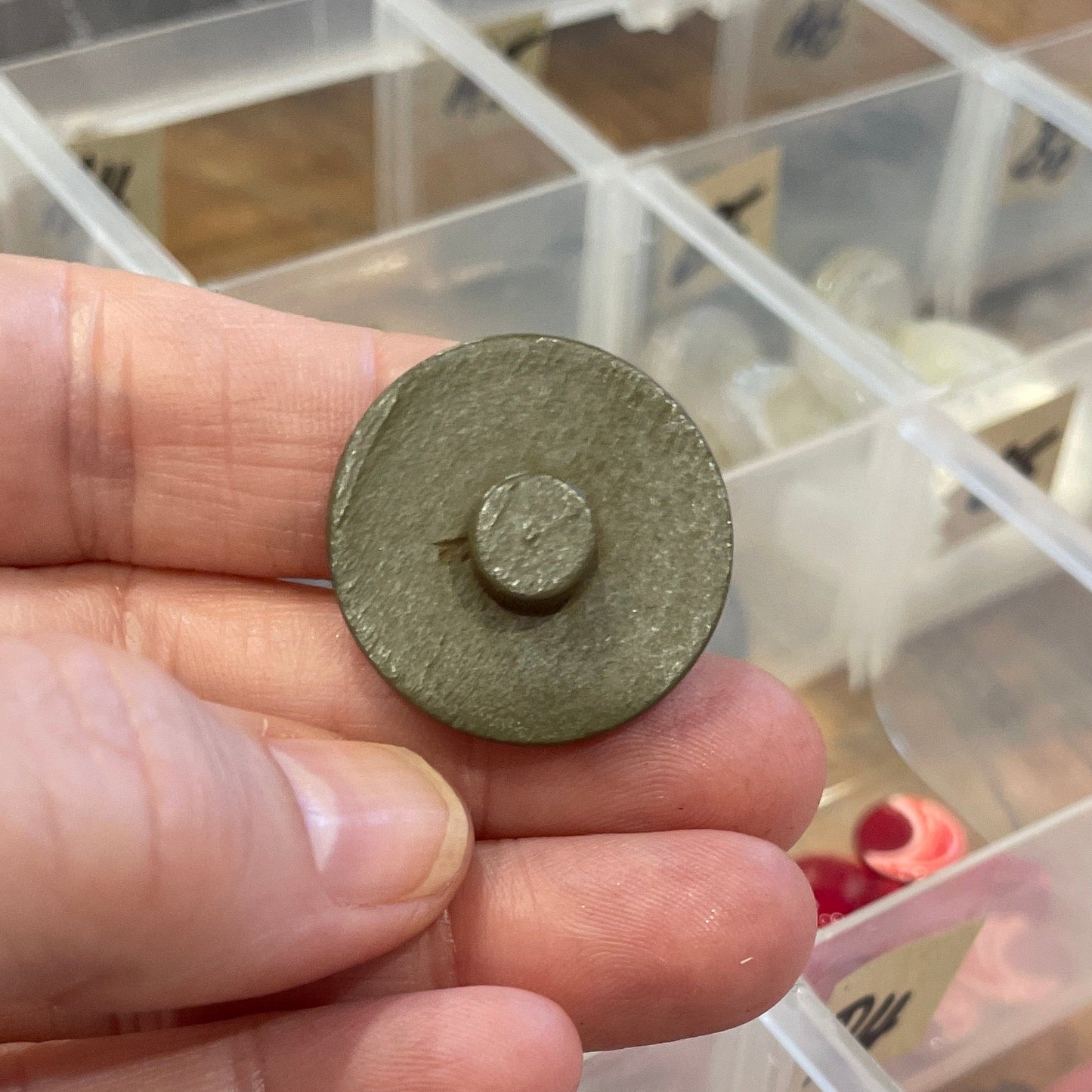 Hand holding a button in front of a clear plastic organizer with various items.
