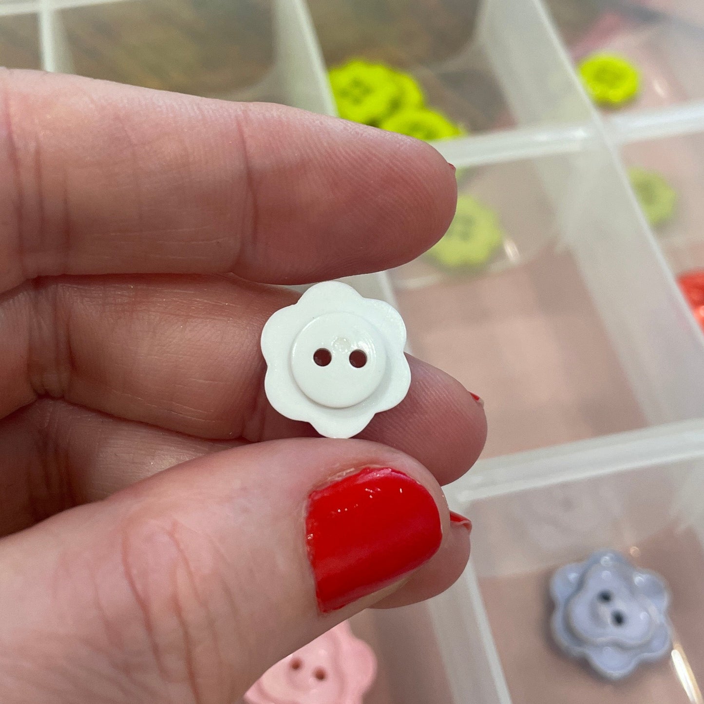 Hand holding a white flower-shaped button with a clear container of buttons in the background