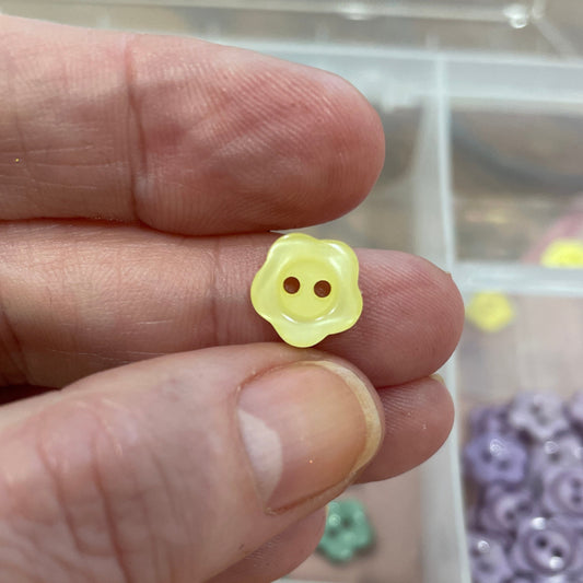 Hand holding a yellow flower-shaped button with a container of buttons in the background