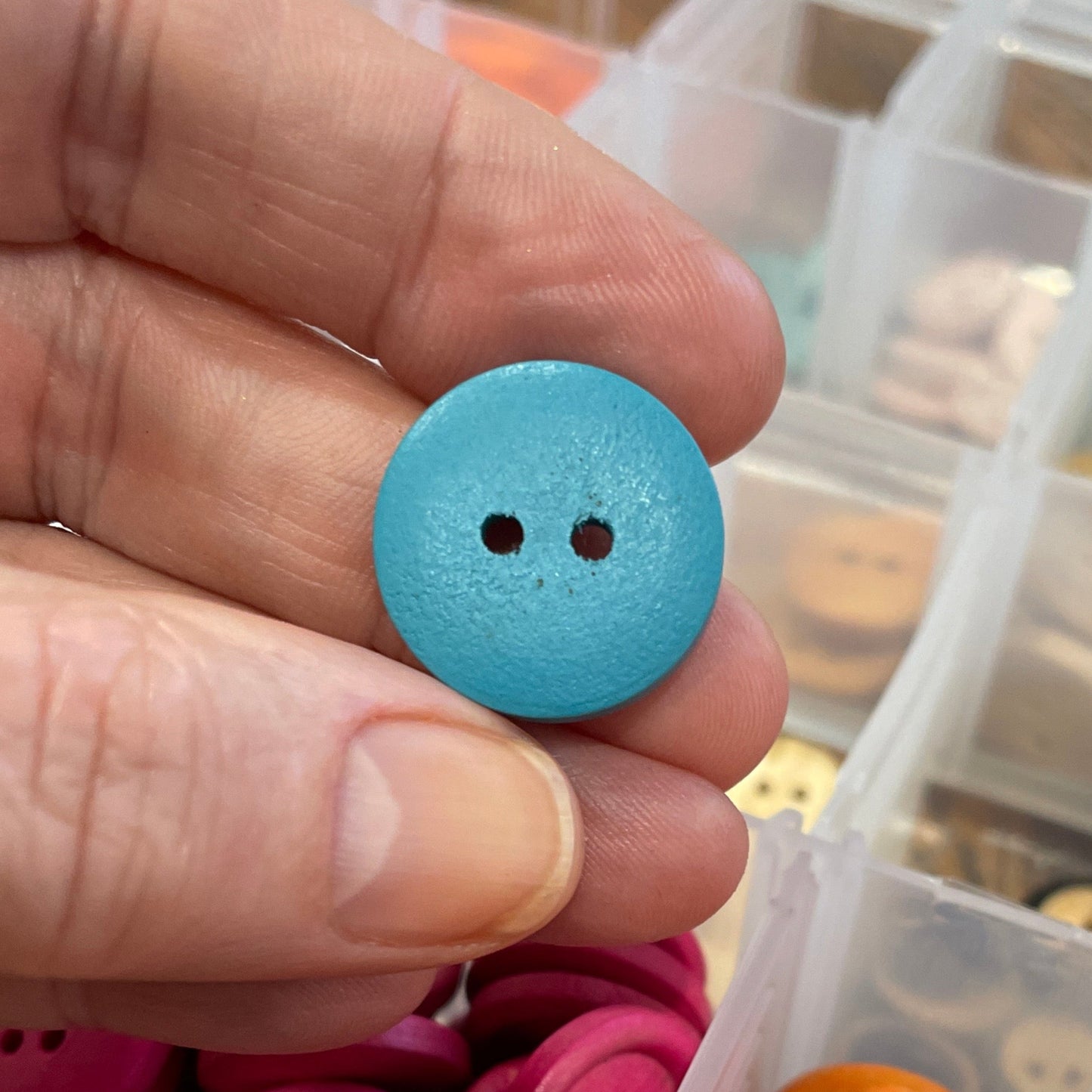 Hand holding a blue button with a container of colorful buttons on a wooden surface