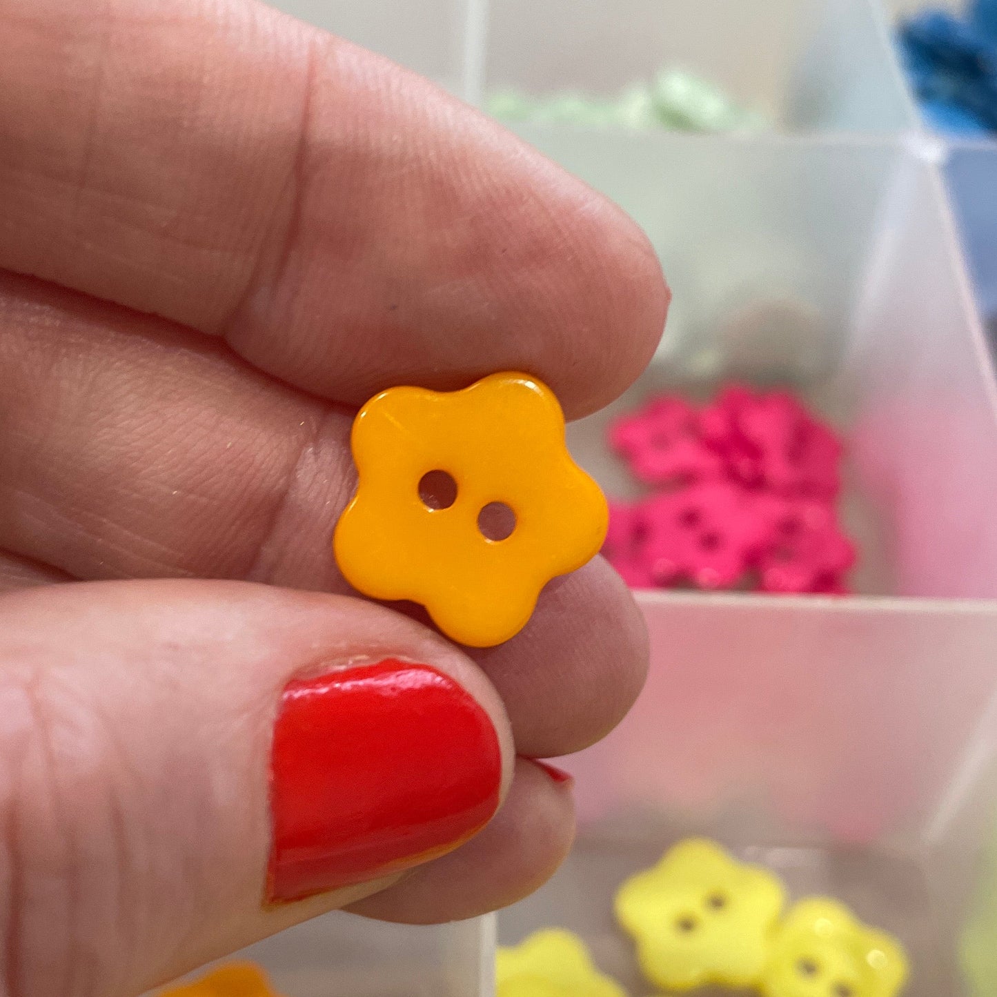 Hand holding a yellow flower-shaped button with a background of more buttons in a container.
