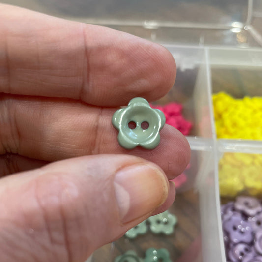 Hand holding a small green flower-shaped button with a clear container of assorted buttons in the background.