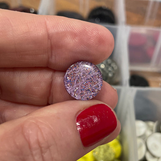 Hand holding a glittery purple button with a container of buttons in the background