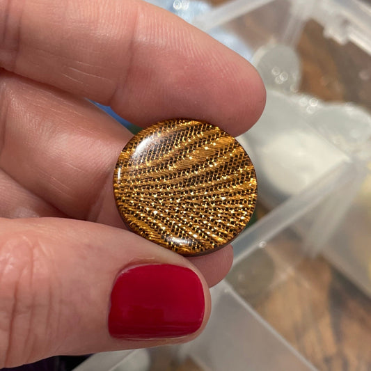 Gold circular object held by a hand with red nail polish, against a background of assorted small items in a clear container.