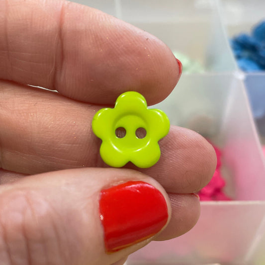 Hand holding a yellow flower-shaped button with a clear container of assorted buttons in the background.