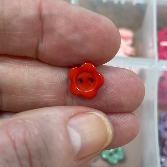 Hand holding a small orange flower-shaped object with a container of assorted buttons in the background.