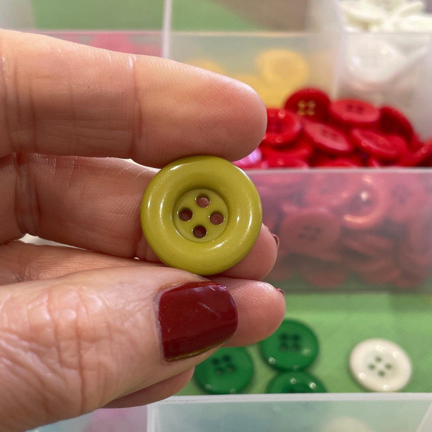 Hand holding a green button with a clear container of assorted buttons in the background