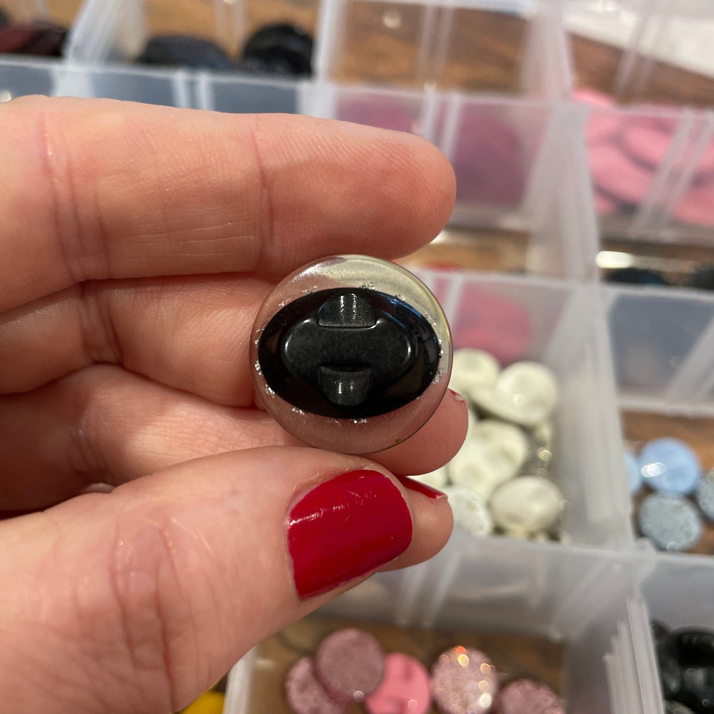 Close-up of a hand holding a small round object with a black center, against a background of colorful buttons in a container.