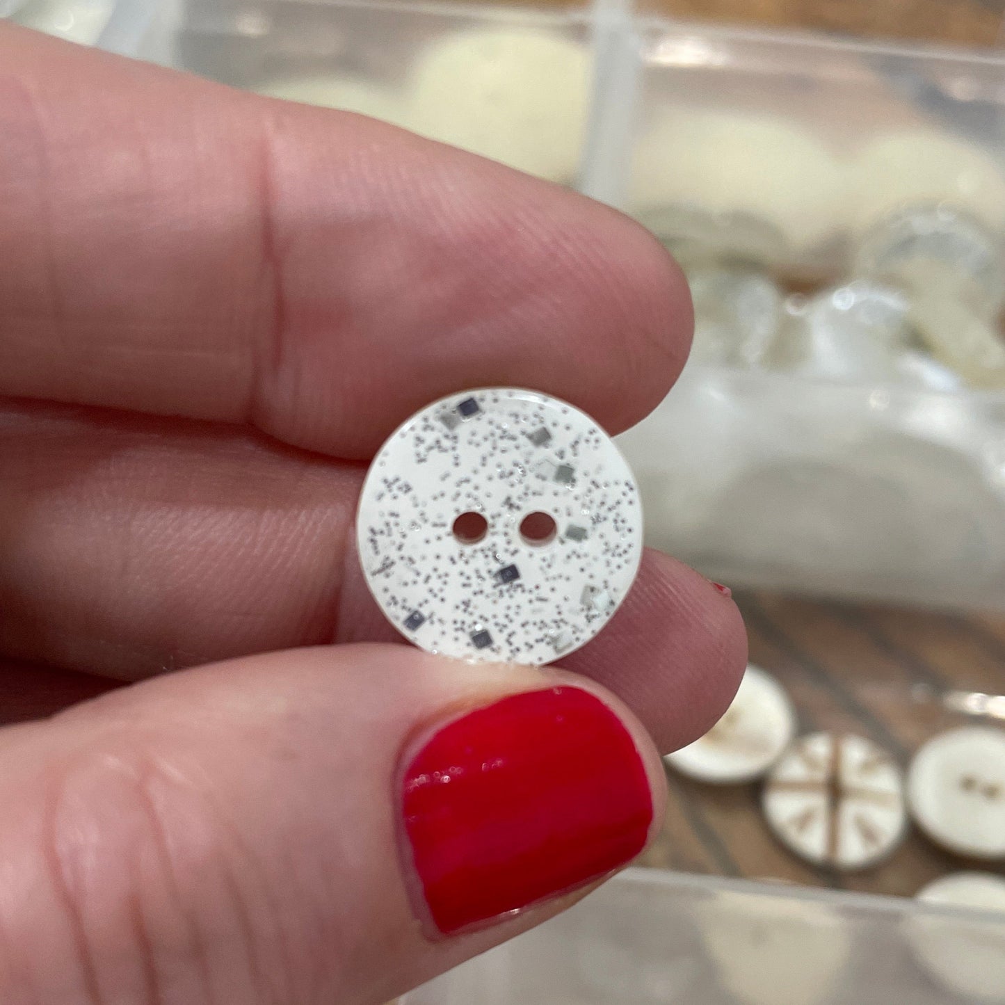 Hand holding a round, glittery button with a clear container of buttons in the background