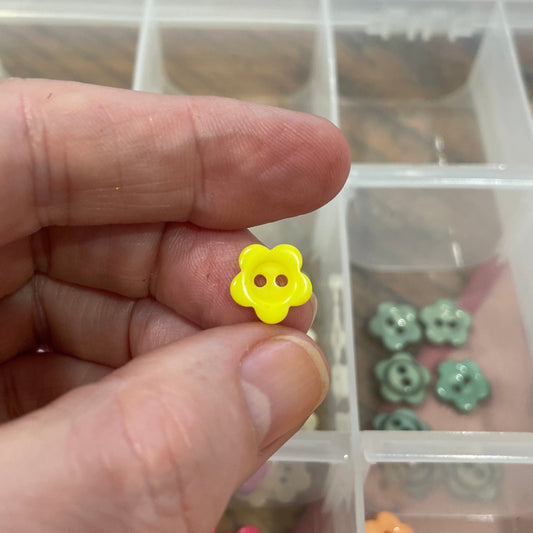 Hand holding a yellow flower-shaped object with a clear container of assorted small objects in the background.