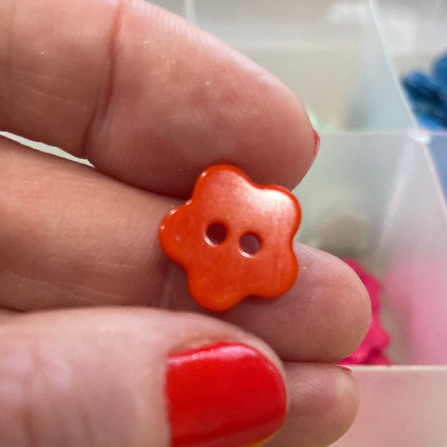 Hand holding an orange flower-shaped button with a container of colorful buttons in the background.