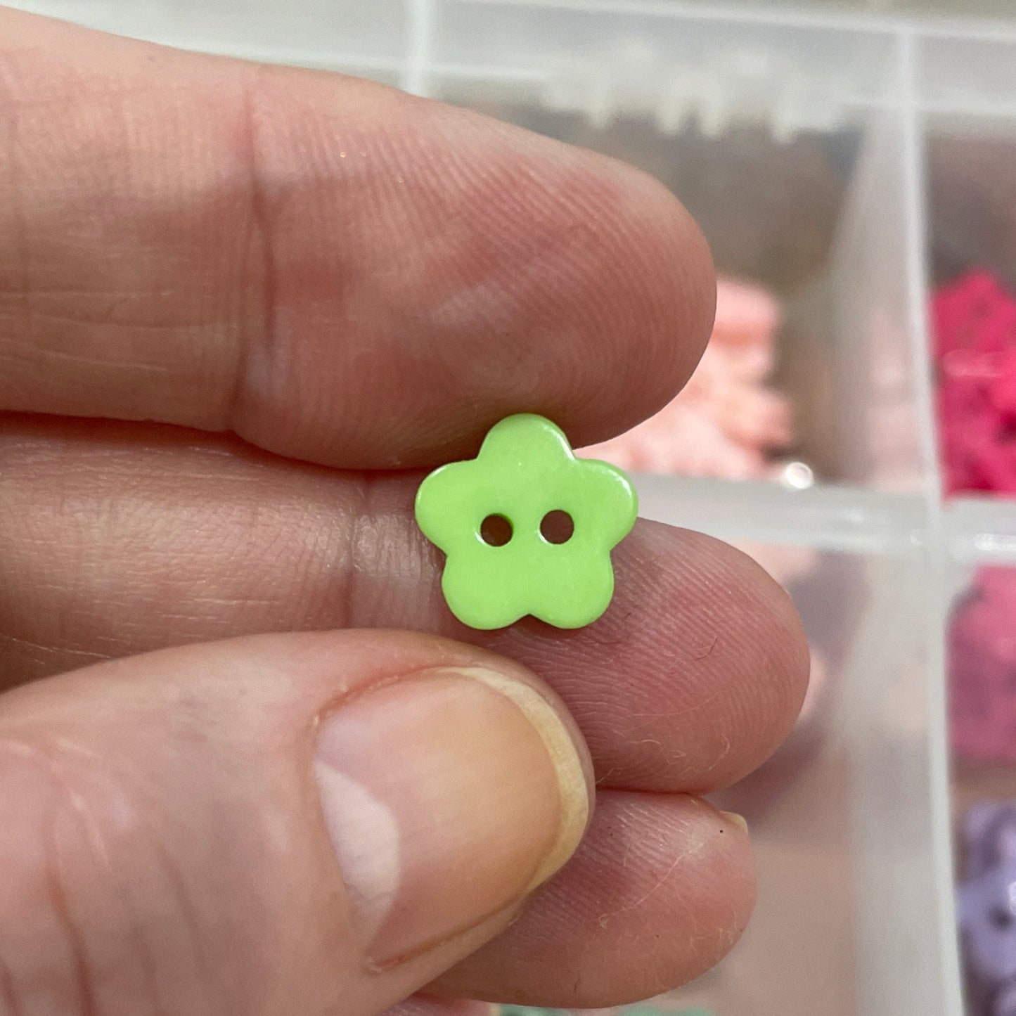 Hand holding a green flower-shaped button with a container of assorted buttons in the background.