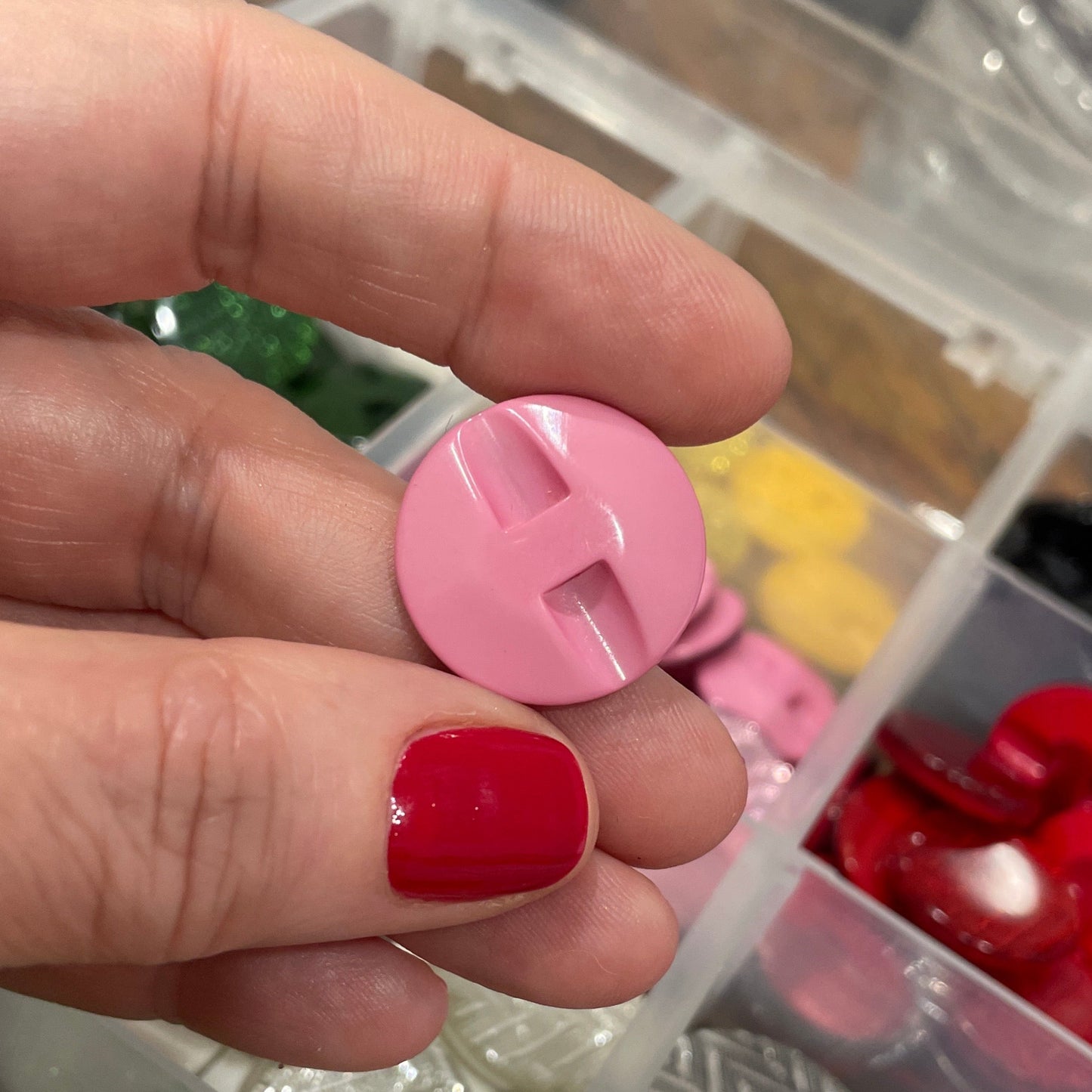 Hand holding a pink button with a container of various buttons in the background
