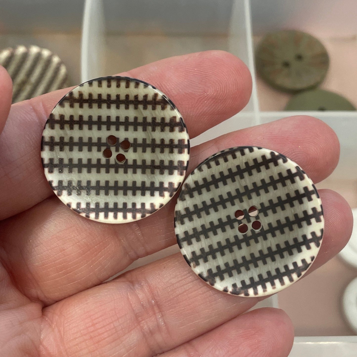 Hand holding two round buttons with a checkered pattern in front of a clear container with more buttons.