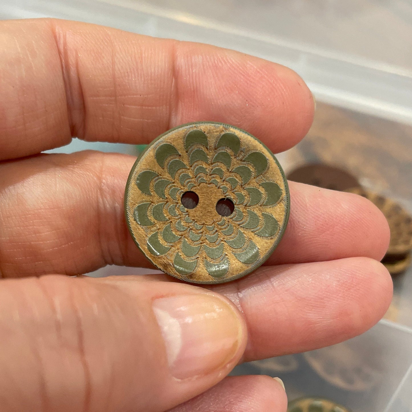 Hand holding a decorative button with a detailed design, against a background of similar buttons in a container.
