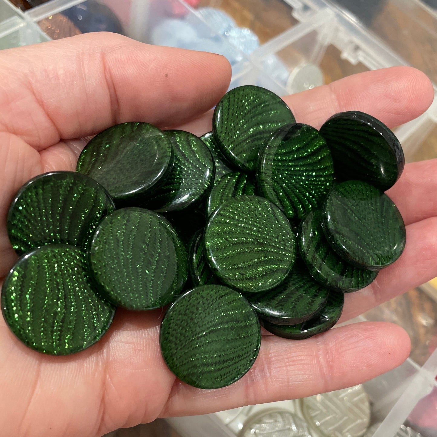 Hand holding green circular objects with a container of various buttons in the background