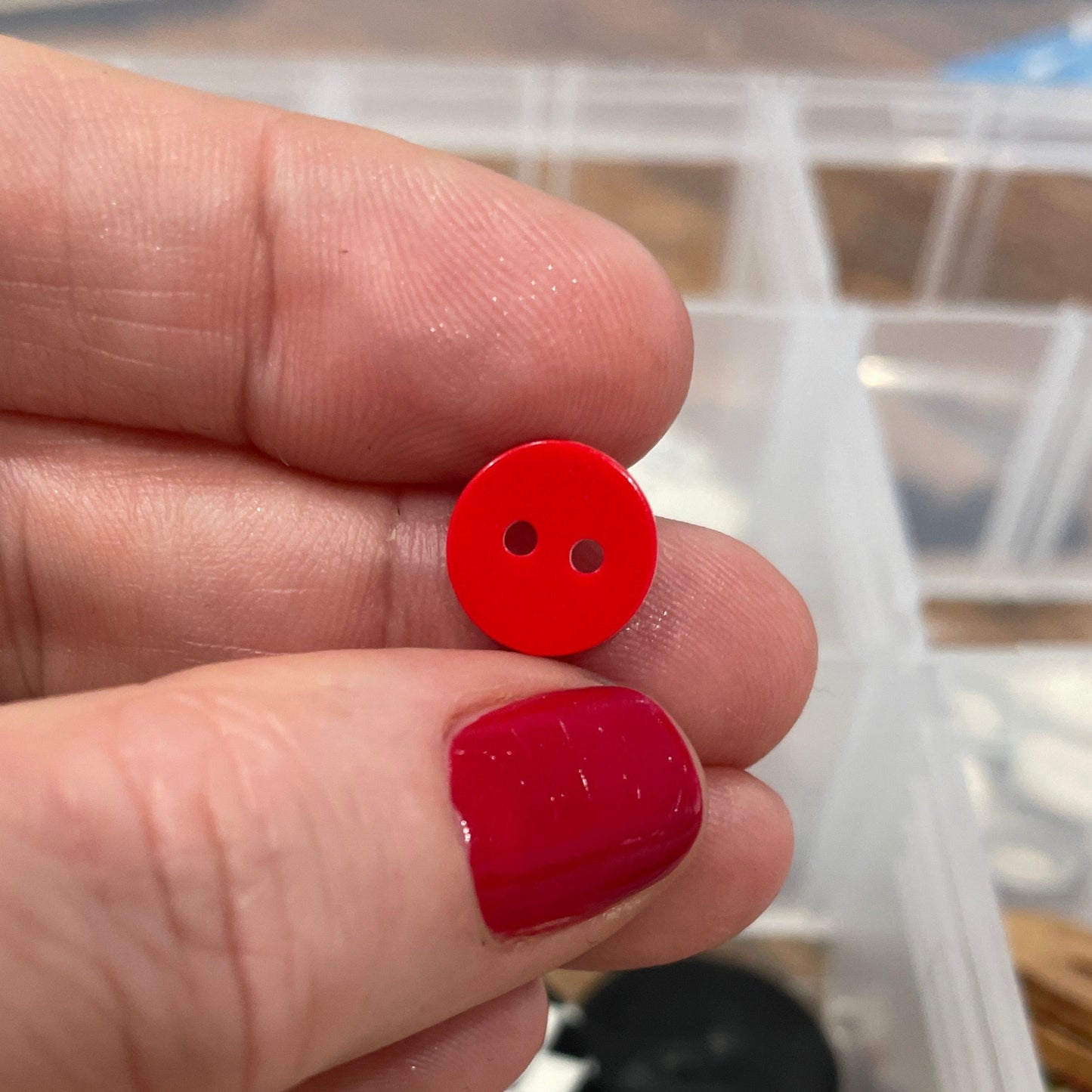 Red button earrings held between fingers with a container of buttons in the background