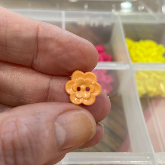 Hand holding a small yellow flower-shaped object with a clear container of assorted small objects in the background.