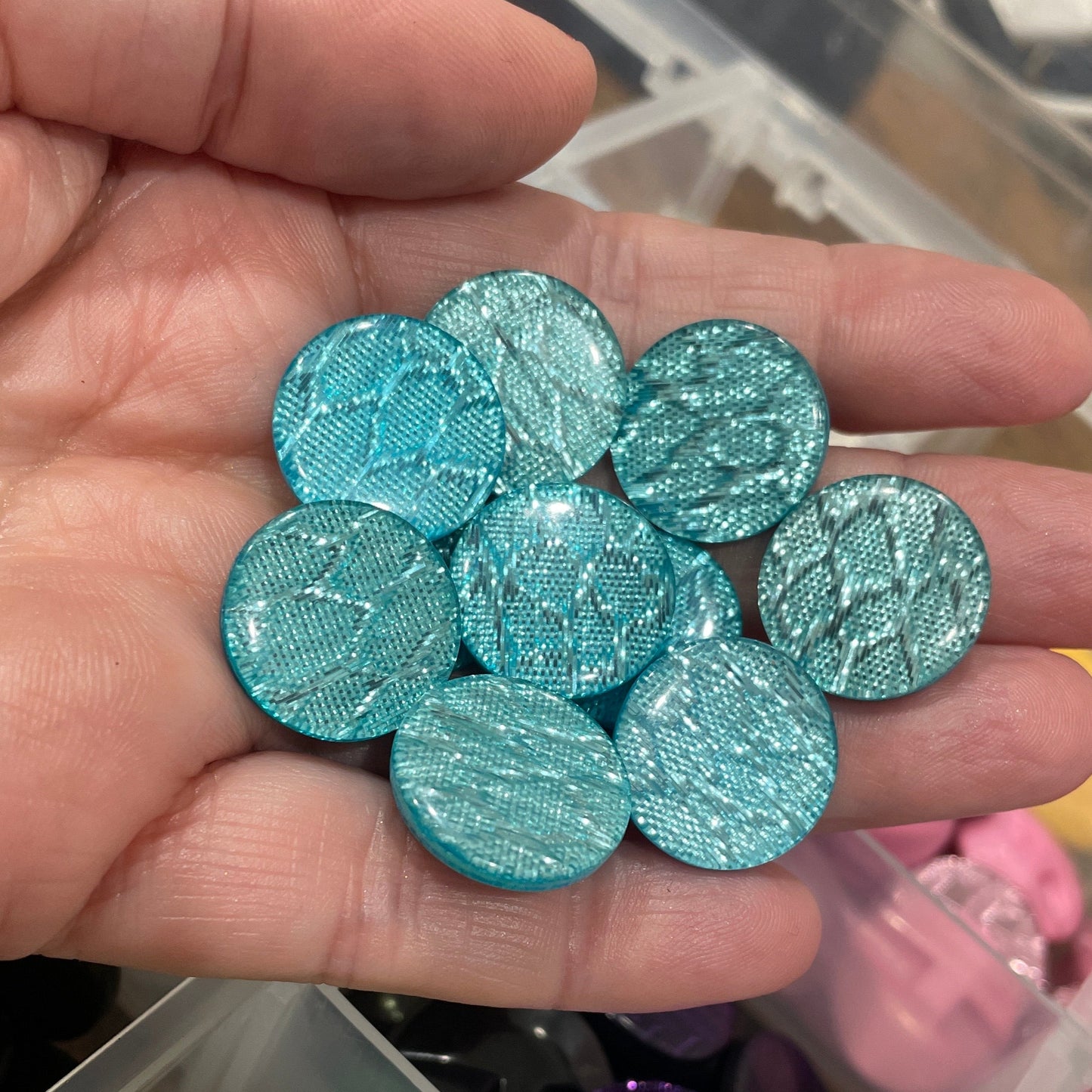 Hand holding teal glittery buttons with a container of colorful buttons in the background.