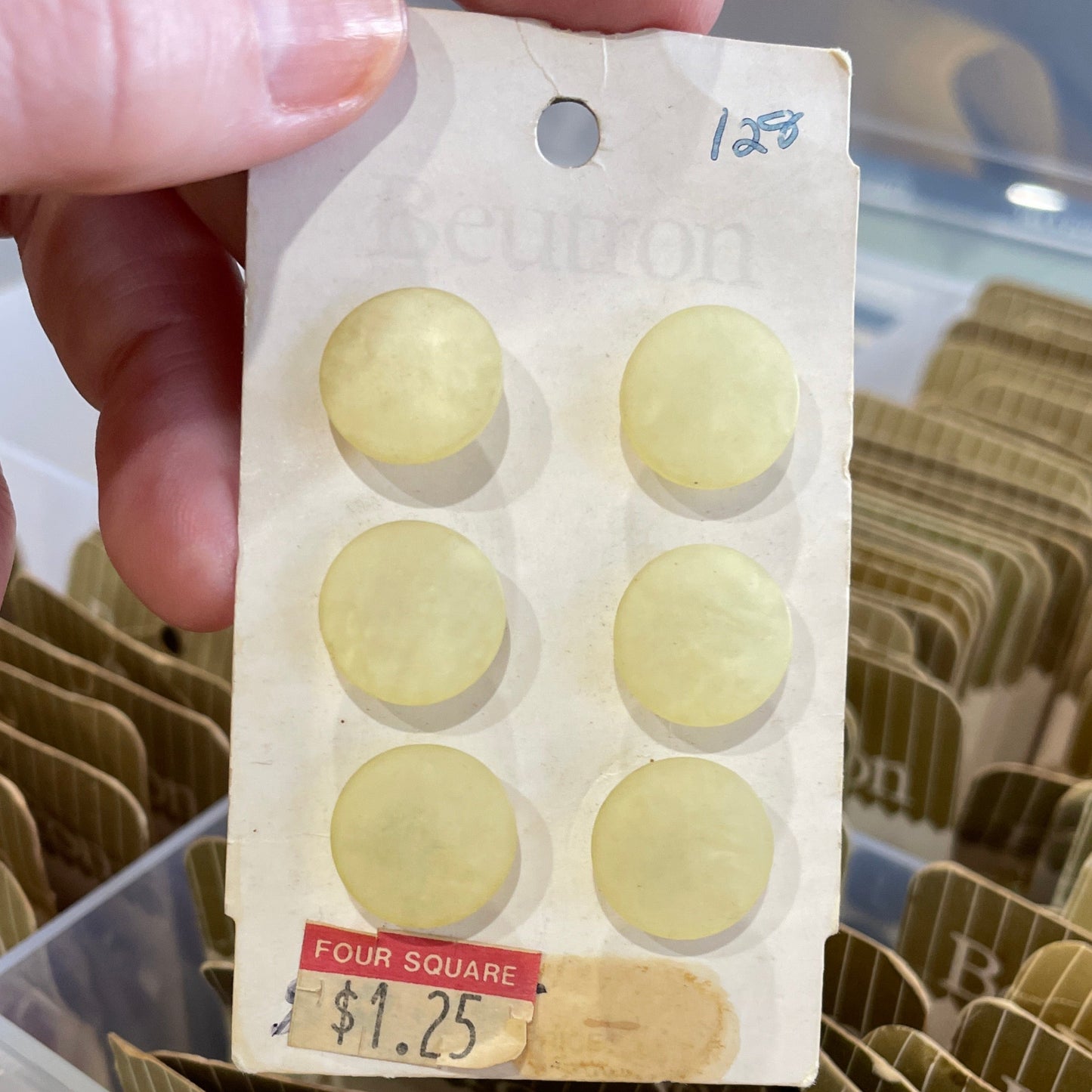 Hand holding a small container with six round beige buttons \, with more containers in the background.