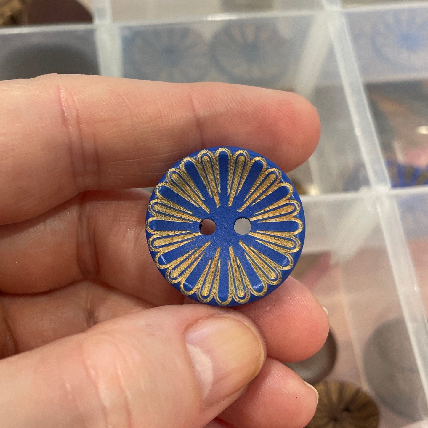 Hand holding a decorative button with a blue and gold design, with a container of buttons in the background.