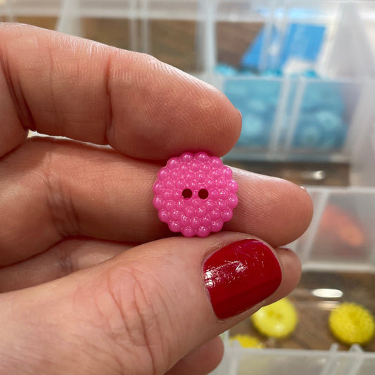 Hand holding a pink button with a blurred background of button storage containers.