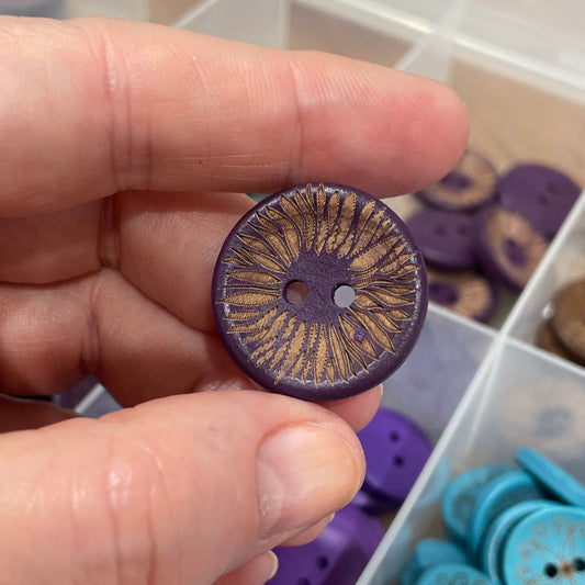Hand holding a purple button with a detailed design, against a background of organized buttons in a container.