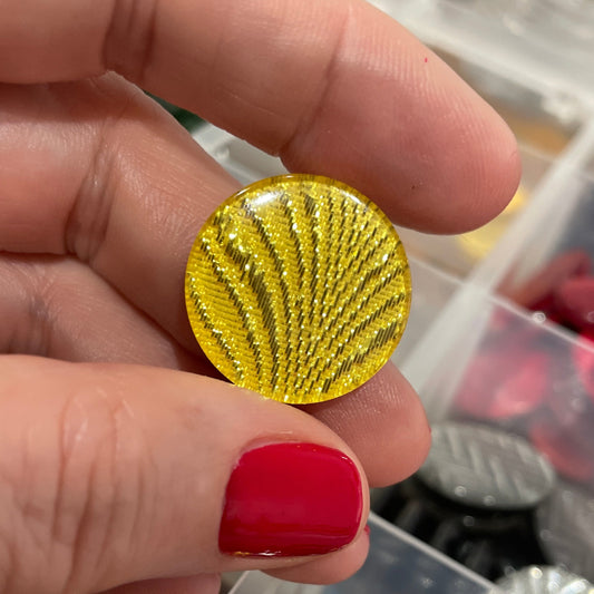 Gold button held by a hand with red nail polish, with a container of colorful buttons in the background.