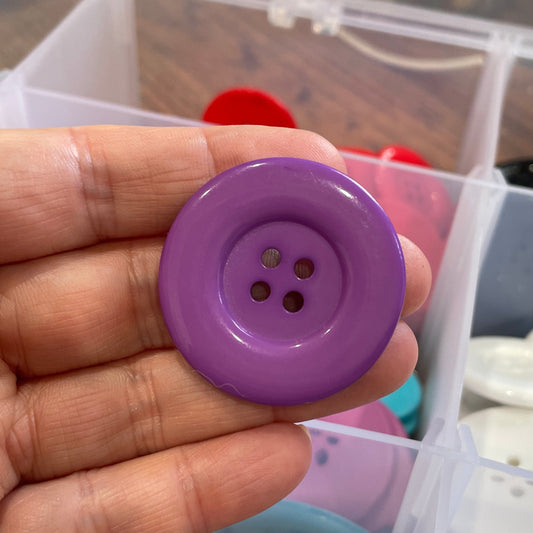 Hand holding a purple button with a container of buttons in the background
