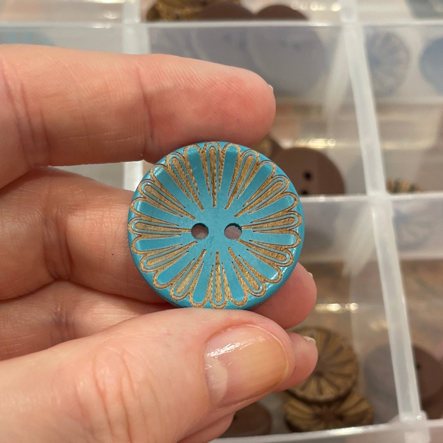 Hand holding a decorative button with a blue and gold design, against a background of a button collection in a box.