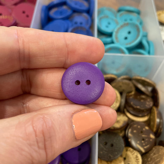 Hand holding a purple button with a container of assorted buttons in the background