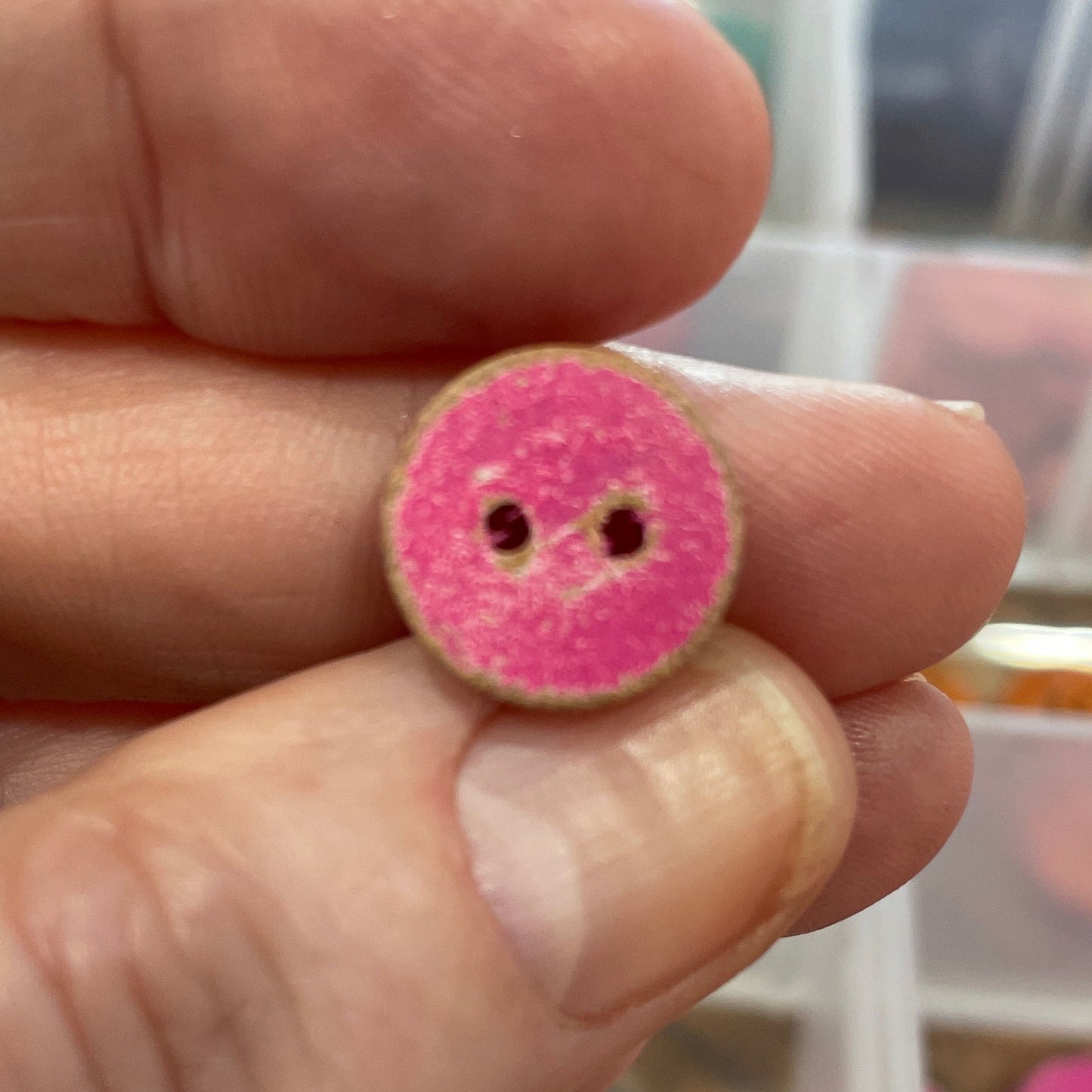 Hand holding a pink button with a blurred background of a button container.