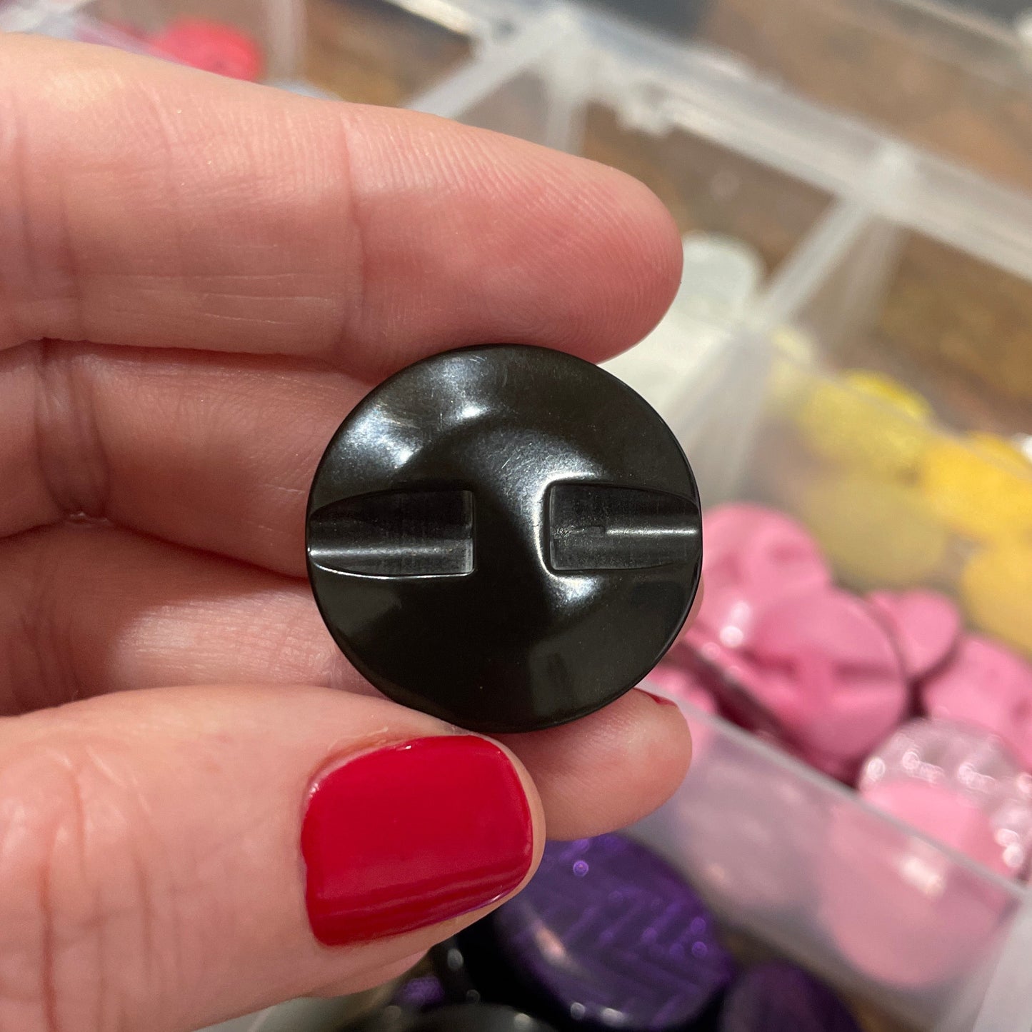 Black plastic component held by a hand with red nail polish, with a colorful assortment of buttons in the background.