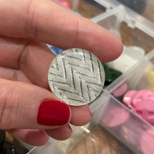 Hand holding a silver button with a textured pattern, in front of a container with various buttons.