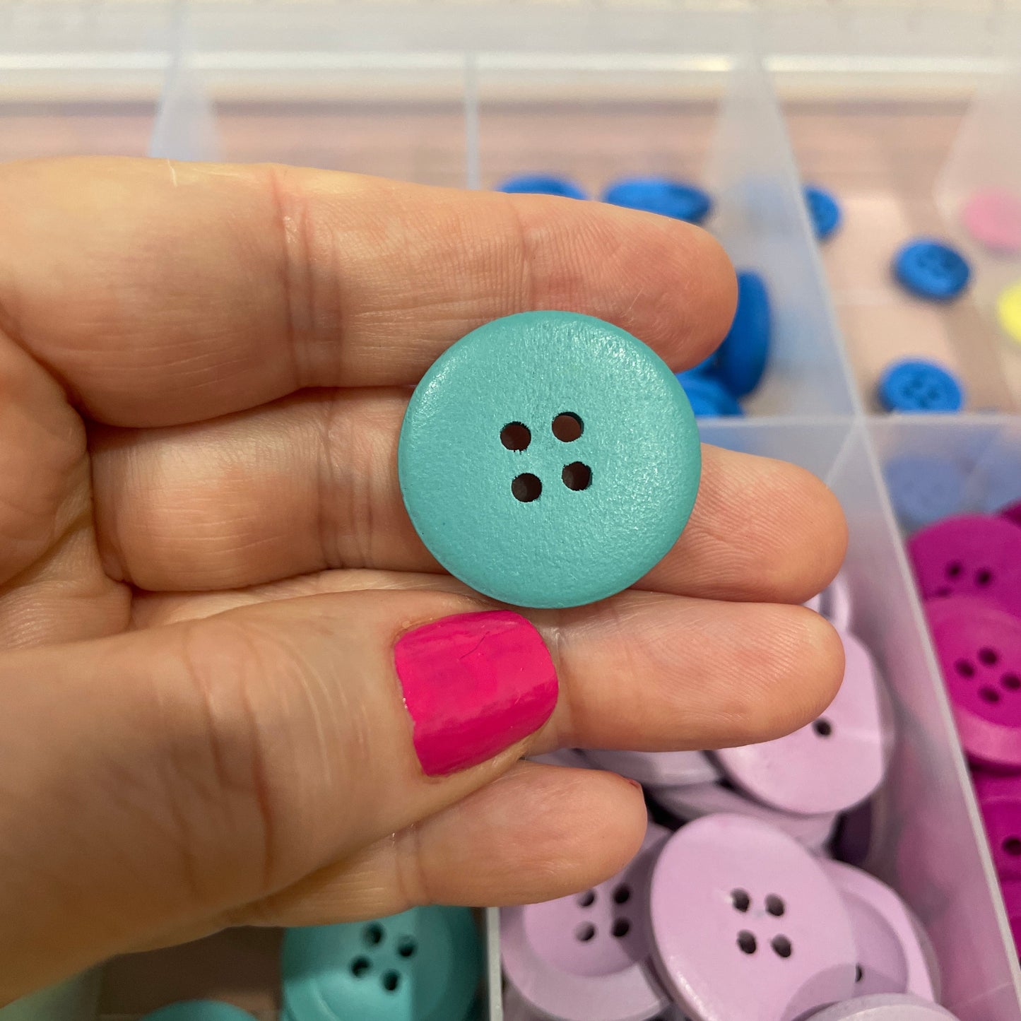 Hand holding a light blue button with a container of colorful buttons in the background